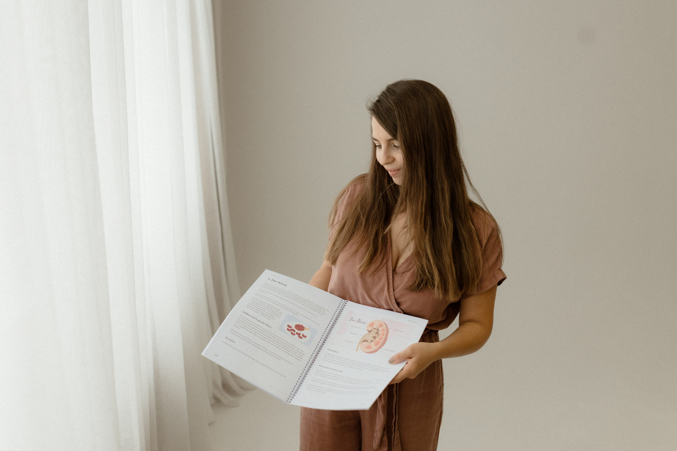 Frau hält Schulungsunterlagen für Fernstudium für die Akademie der Naturheilkunde in der Hand und präsentiert sie neben einem weißen Vorhang