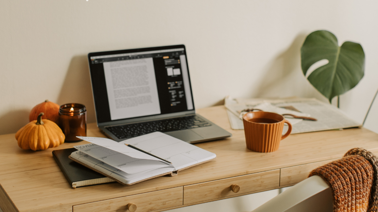 Schreibtisch als Arbeitsplatz mit Laptop, Notizbuch, brauner Tasse, Monstera Pflanze nebendran und Kürbissen als Deko.