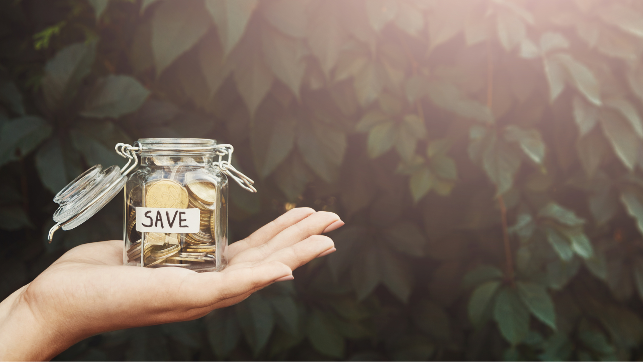 Weibliche Hand hält Spargläschen mit Cent drin. Die Nägel sind manikürt und vor einem Hintergrund, der aussieht, wie eine Hecke.